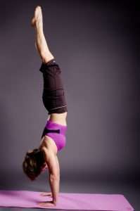 A person in a purple top and black pants performs a handstand on a pink yoga mat against a dark background, embodying the balance and focus of a plant-based lifestyle. Their legs extend straight up, showcasing strength and dedication.