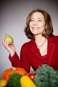 A person in a red blazer, reminiscent of a cheerful dietitian, smiles while holding a pear. In front of them are assorted fruits and vegetables, including a pumpkin, squash, and leafy greens. The softly blurred background complements this vibrant vegetarian tableau.