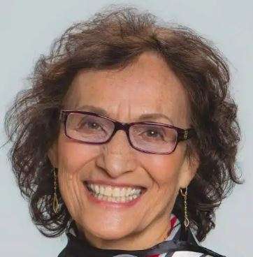 Smiling older adult with glasses, short curly brown hair, and wearing earrings, exuding a sense of wisdom against a light background. As an advocate for a plant-based lifestyle, they embody the vibrant spirit of a dedicated dietitian.