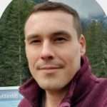 Portrait of a person with short hair wearing a maroon jacket. They stand outdoors, surrounded by evergreen trees and a cloudy sky, embodying the serene lifestyle of a vegetarian enthusiast.