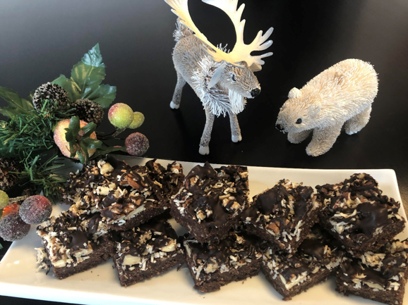 A plate of decadent chocolate brownies topped with nuts and coconut sits invitingly on the table. Beside it, whimsical figures of a reindeer and polar bear add charm, while frosted berries and greenery complete this delightful vegan-friendly display.