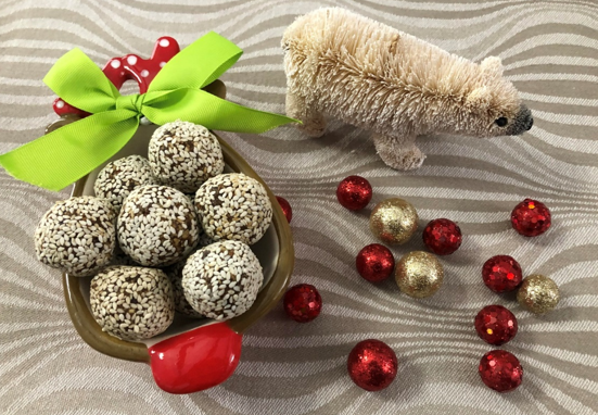 A ceramic sleigh brimming with sesame-covered treats is adorned with a green bow, offering a delightful plant-based option. Beside it sits a plush polar bear and scattered red and gold glittery baubles on wavy-patterned fabric.