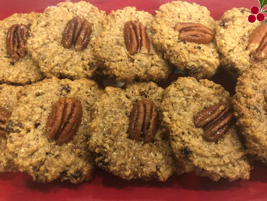 A close-up of a batch of homemade, plant-based cookies arranged on a red surface. Each cookie is topped with a whole pecan, showcasing a crumbly texture with visible grains and seeds baked into them—perfect for those embracing a vegetarian lifestyle.