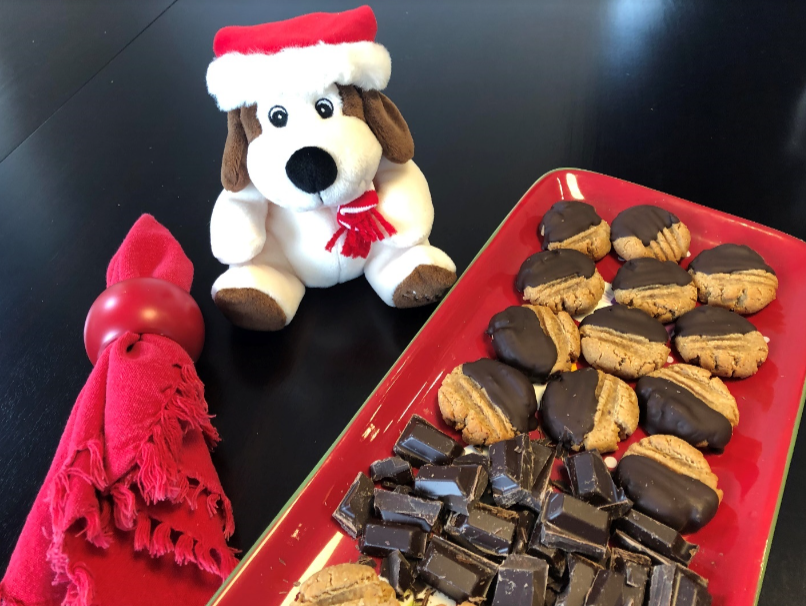 A festive stuffed toy wearing a Santa hat and red scarf sits beside a red napkin with a ring on the table. A platter holds plant-based peanut butter cookies partly dipped in chocolate and a pile of chocolate pieces.