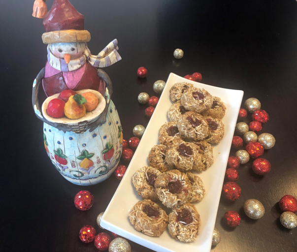 A ceramic snowman figurine holding fruit stands next to a rectangular white plate of vegan cookies topped with jam and coconut. Red and gold glittery ornaments are scattered on the dark surface beneath them, adding a festive touch.