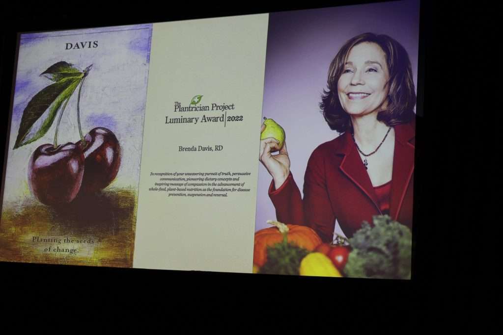 A screen displays an award presentation slide. On the left is a painting of cherries with leaves. The middle text reads The Plantrician Project Luminary Award 2022. On the right, a smiling dietitian in a red jacket holds an apple among plant-based vegetables.