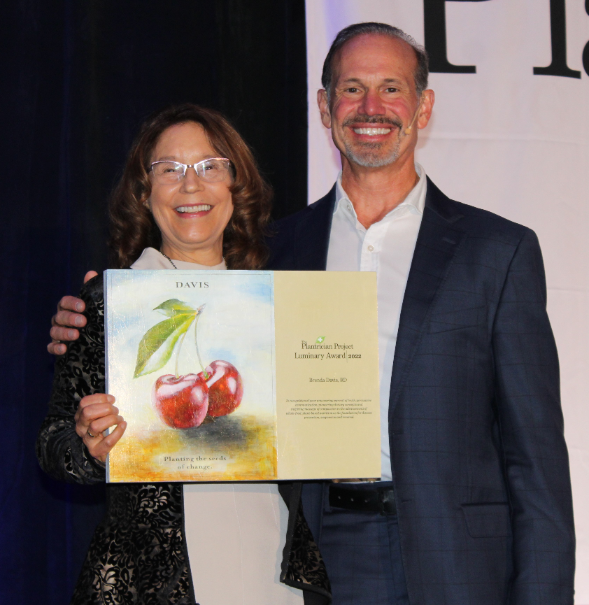 A woman and a man are smiling and standing together. The woman, a renowned dietitian, holds an award depicting apples with the text Planting the seeds of change. Plant Pure Project Luminary Award 2022 Brenda Davis, RD. Both are dressed in formal attire, celebrating achievements in plant-based nutrition.