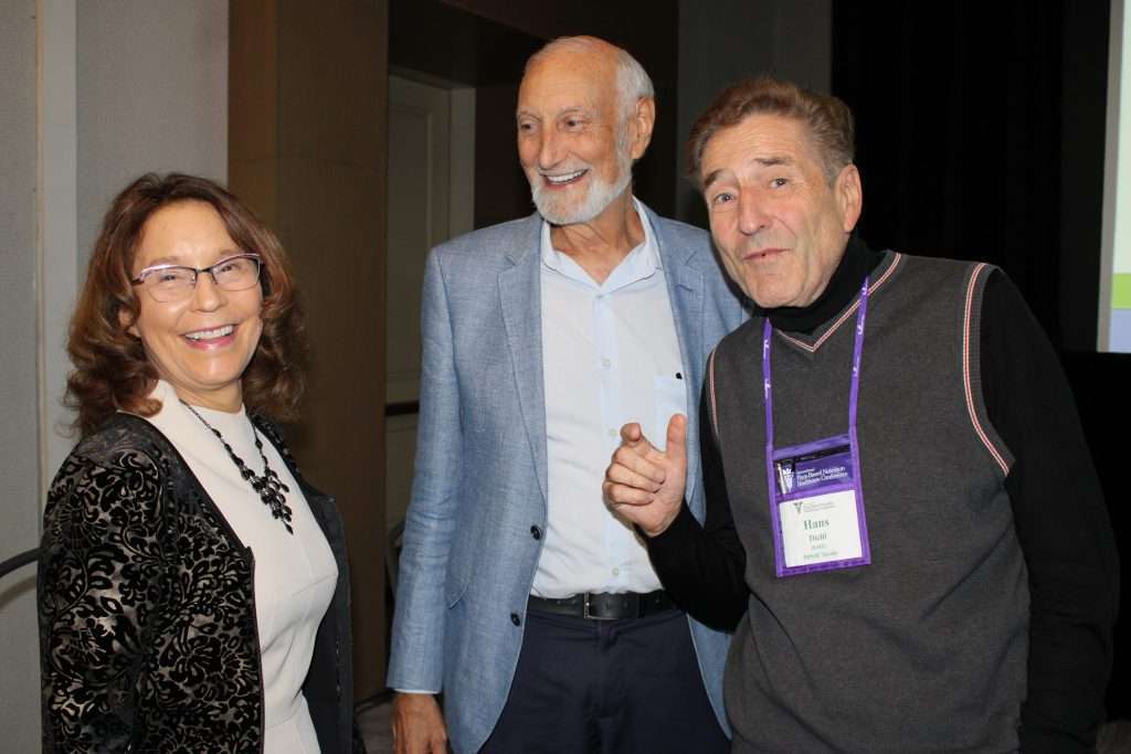 Three people are smiling and talking at an event. The person on the left, a dietitian, is wearing glasses and a black-and-white outfit. The person in the middle sports a light blue jacket, while the one on the right, wearing a name tag, gestures enthusiastically about plant-based choices.