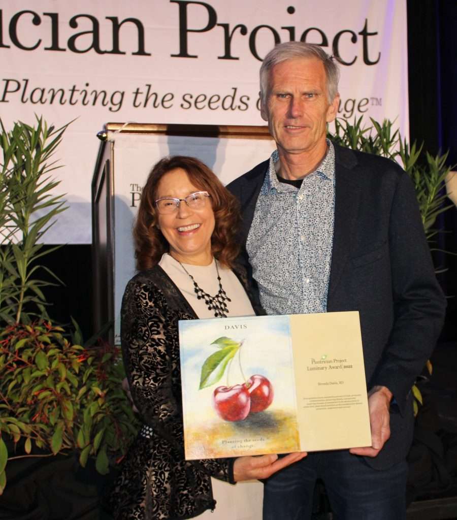 A woman and a man stand together smiling. The woman, a dietitian, holds a plaque featuring painted cherries and text. Behind them is a banner that reads, Planting the seeds of change. They appear to be at an event celebrating plant-based initiatives.