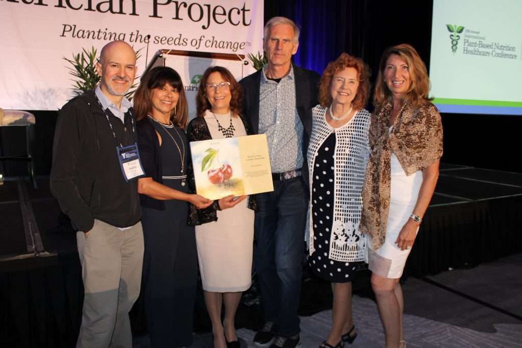A group of six people stands on a stage holding a book. They are smiling at the camera, dressed in business casual attire. A banner in the background reads “Planting the seeds of change” with a logo on the right side, symbolizing their commitment to a plant-based future.