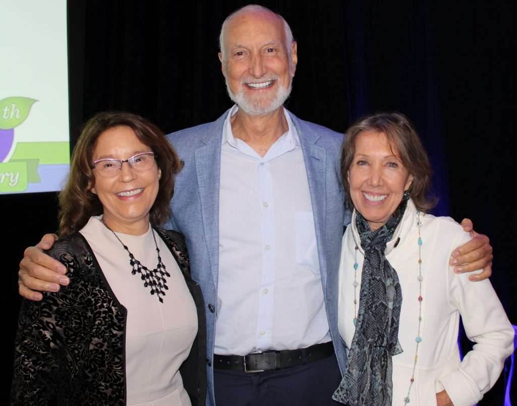 Three people are standing together, smiling at the camera. The man in the middle has a light beard, wearing a light blue suit jacket. The woman on the left, possibly a dietitian, wears glasses and a black necklace, while the woman on the right sports a white jacket and scarf.