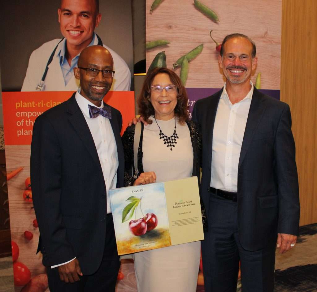 Three people smiling in front of a backdrop with fruit and plant imagery. The woman in the middle, likely a dietitian, holds a certificate adorned with apple illustrations. They are dressed formally, with the men in suits and the woman elegantly attired in a dress and shawl.