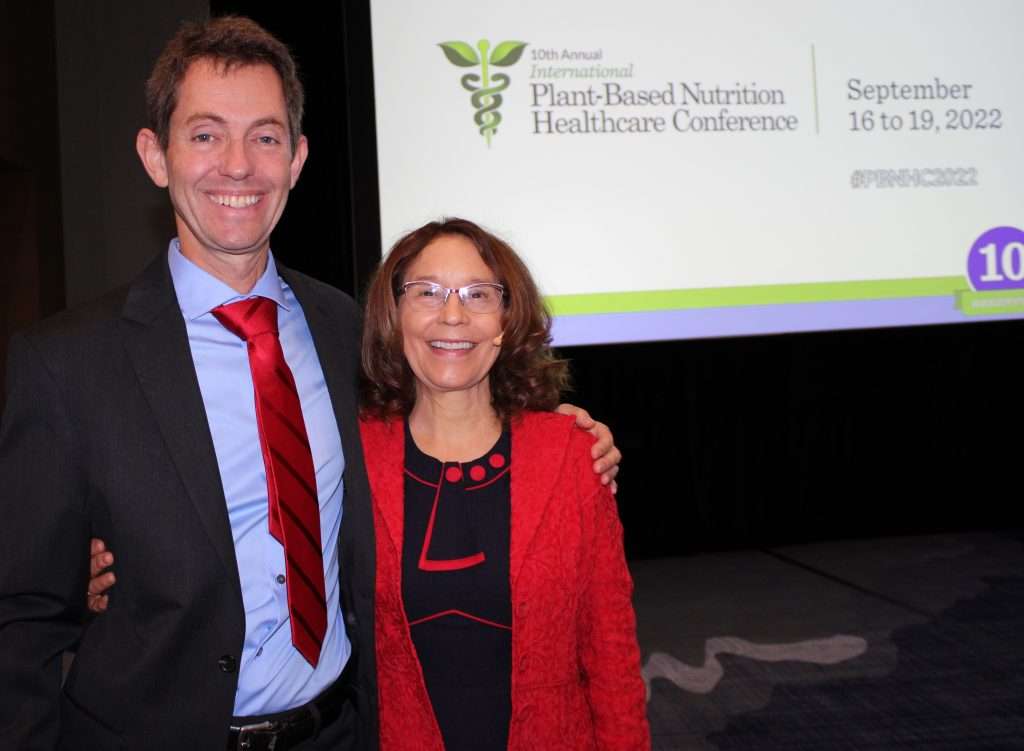 A man and woman, both smiling and posing together at the International Plant-Based Nutrition Healthcare Conference, exemplify the vibrant spirit of vegetarian advocacy. The screen behind them highlights event details: September 16 to 19, 2022. They are both dressed in business attire.
