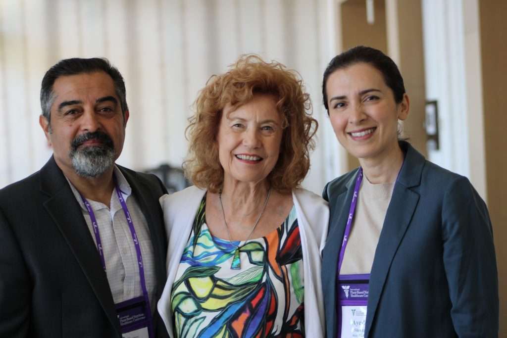 Three people smiling at a professional event, wearing conference name tags. The man on the left, perhaps a plant-based dietitian, has a beard and is in a suit. The woman in the center has curly red hair and a colorful dress. The woman on the right has dark hair pulled back, wearing a blazer.