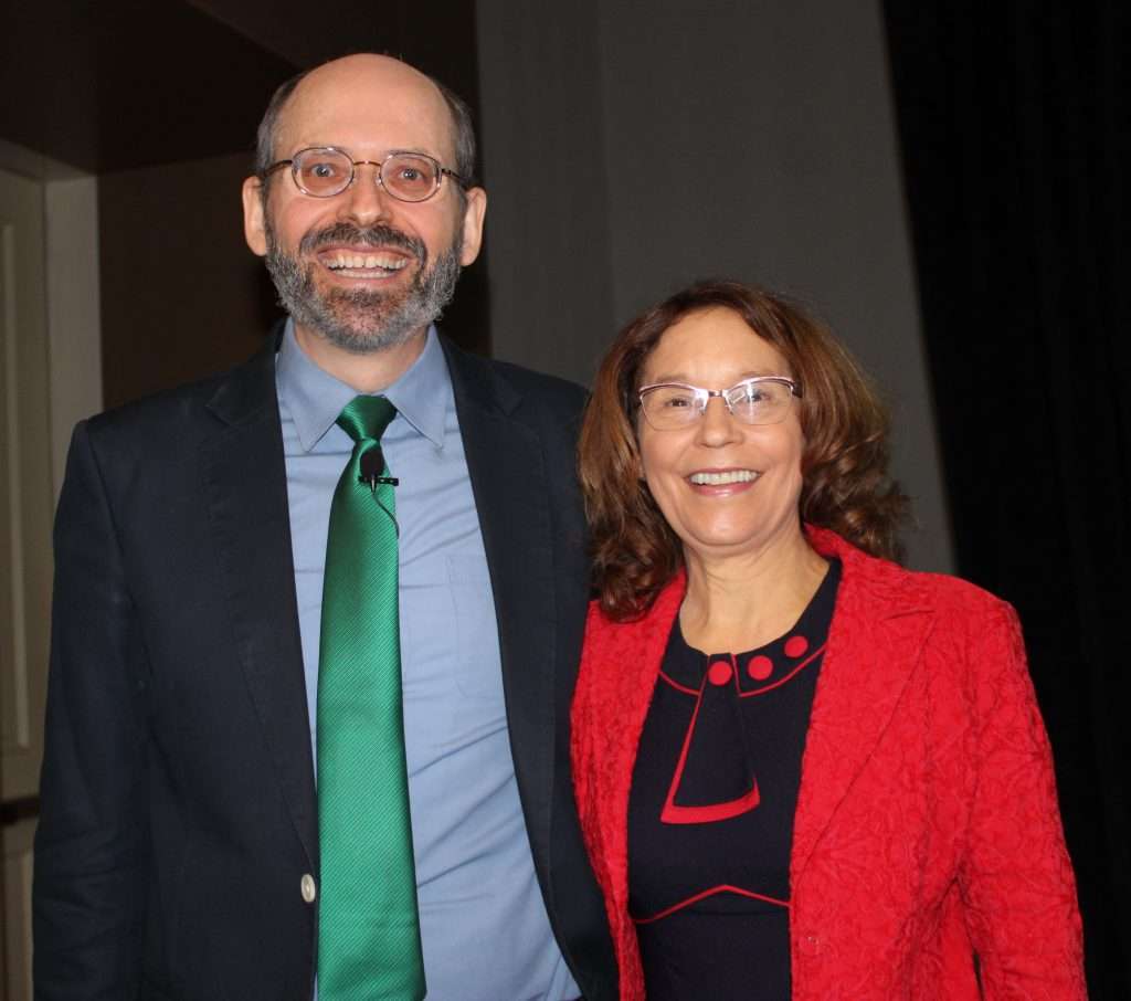 A man and a woman, both radiant with smiles, are captured on camera. The man dons a suit with a green tie, while the woman sports a red jacket over a black top. Perhaps theyre celebrating after consulting their favorite dietitian about the benefits of plant-based living indoors against a dark backdrop.