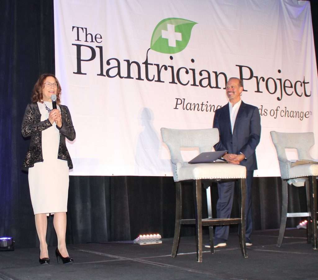 Two people stand on stage beneath a banner reading The Plantrician Project: Planting seeds of change. One speaks into a microphone about vegan lifestyles while the other, possibly a dietitian, is at a high table with a laptop. Two high chairs complete the setup.