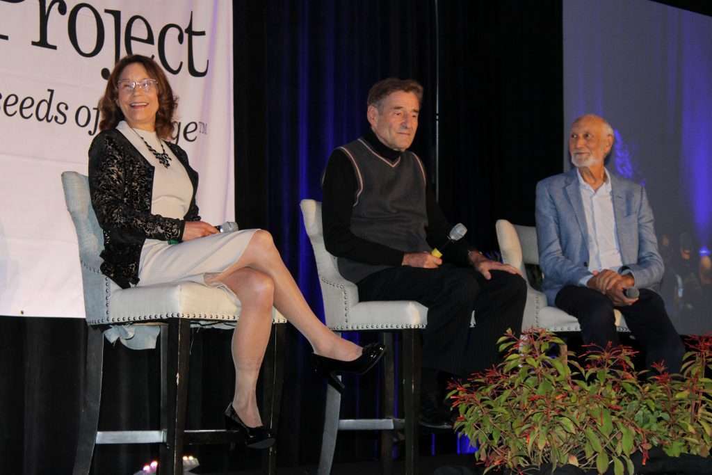 Three people are seated on stage with microphones. The woman on the left, possibly a dietitian, wears a black and white outfit, the man in the middle wears a dark sweater, and the man on the right sports a light blue suit. A banner and lush plants are in the foreground.