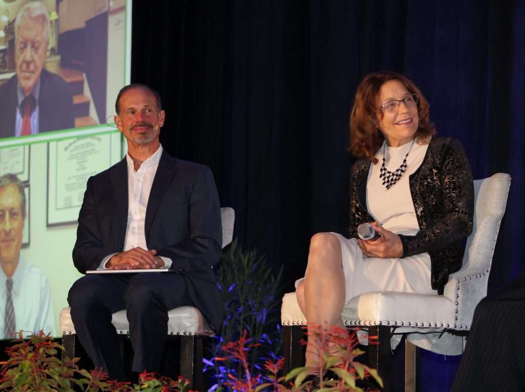 A man in a suit and a woman in a black jacket and white dress, perhaps a dietitian, sit on stage with microphones. Theyre smiling as a screen behind them shows images of other people. A lush array of plants is visible in the foreground, complementing the scenes vibrant energy.