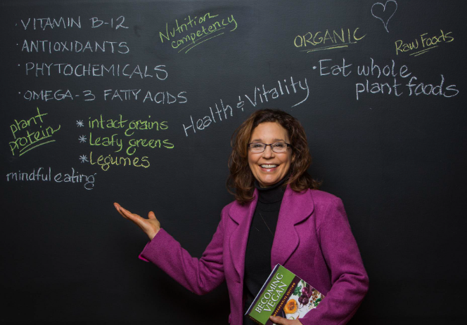 A woman in a purple blazer smiles as she gestures towards a blackboard displaying handwritten notes on nutrition, featuring Vitamin B-12, antioxidants, and plant protein. As a dietitian, she holds the book Becoming Vegan, emphasizing her passion for plant-based living.