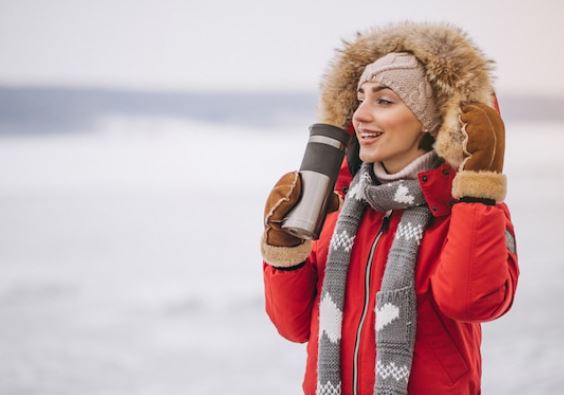 A person stands outdoors in a snowy landscape, wearing a red winter coat with a fur-lined hood, a knitted hat, and gloves. They hold a travel mug filled with a vegan hot chocolate blend and appear to be smiling. The background is blurred with snow.
