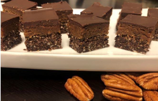 A plate with several chocolate layered desserts featuring a crunchy base, a middle layer, and a smooth top sits enticingly. Perfect for vegetarians, pecans are scattered below the plate on a dark surface.