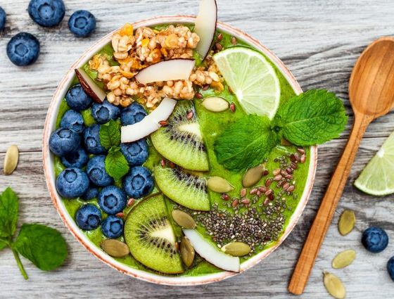 A vibrant, plant-based smoothie bowl on a wooden surface, topped with kiwi slices, blueberries, coconut flakes, pumpkin seeds, chia seeds, granola, lime slices, and mint leaves. A wooden spoon and a lime wedge are beside the bowl—an ideal creation for any dietitians vegetarian delight.