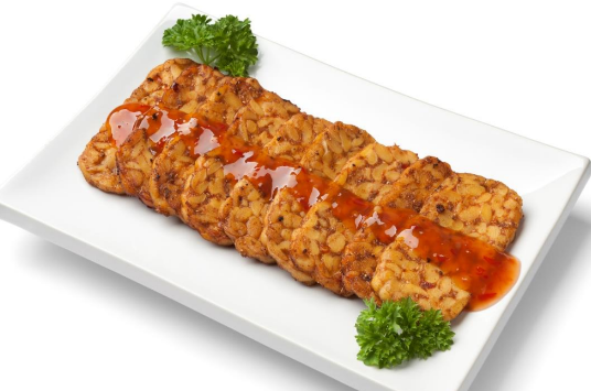 Slices of fried tempeh drizzled with a red sweet chili sauce are neatly arranged on a white rectangular plate, showcasing a delectable plant-based dish. Several sprigs of fresh parsley garnish the corners, adding both color and flavor.