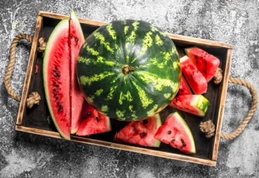 A wooden tray holds a whole watermelon and several slices, perfect for a refreshing vegan treat. The watermelon features a dark green rind with light stripes. Juicy red flesh dotted with black seeds is revealed in the slices, all resting on a textured gray surface.