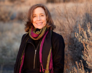 A smiling woman with shoulder-length brown hair stands outdoors in a natural setting, wearing a black coat and a colorful scarf. As a dietitian with a plant-based lifestyle, she enjoys the dry grass and soft, warm lighting of natures embrace.