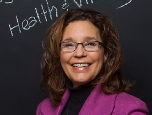 A smiling woman with wavy brown hair and glasses stands in front of a chalkboard. Shes wearing a purple jacket and a black turtleneck. The chalkboard has partial writing that includes the words Health & visible.