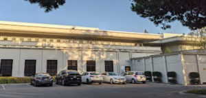 A parking lot with six cars is in front of a white building with large windows. The building has a flat roof and is surrounded by trees. Shadows from the trees fall on the buildings wall and the pavement. The sky is clear.