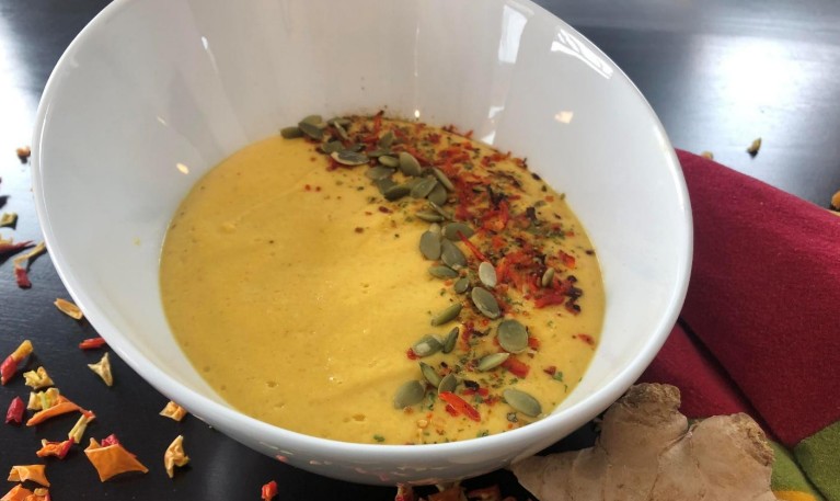 A white bowl filled with creamy yellow soup, garnished with pumpkin seeds, red pepper flakes, and herbs, sits on a dark table next to a piece of ginger and a red napkin.