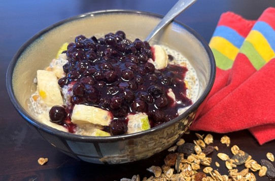 A bowl of chia pudding topped with banana slices and blueberry compote, with a spoon in the bowl. A colorful napkin and scattered granola are next to the bowl on a dark surface.