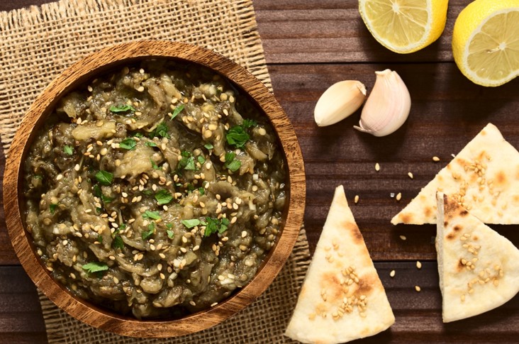 A wooden bowl of baba ganoush garnished with parsley and sesame seeds sits on a burlap mat. Nearby are pita triangles, garlic cloves, and halved lemons on a wooden table.