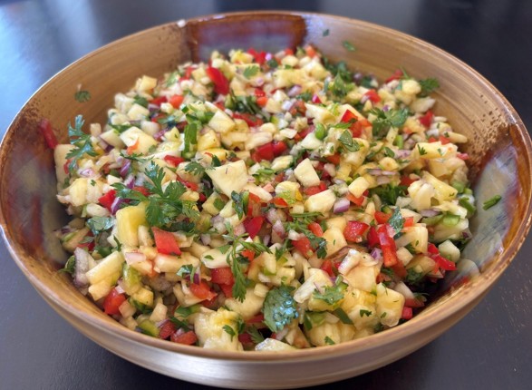 A wooden bowl filled with a colorful chopped salad containing pineapple, red bell pepper, onions, cilantro, and other fresh ingredients, placed on a dark table.