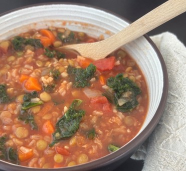 A bowl of vegetable soup with kale, carrots, lentils, rice, and tomatoes, with a wooden spoon resting inside and a beige napkin beside the bowl.