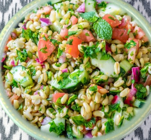 A close-up of a bowl filled with tabbouleh salad, featuring bulgur wheat, chopped tomatoes, cucumbers, red onions, parsley, and mint on a patterned cloth background.