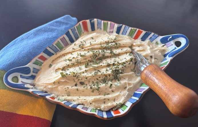 A colorful dish holds a creamy spread topped with herbs, accompanied by a wooden-handled spreader. A blue cloth napkin sits beside the dish on a dark tabletop.