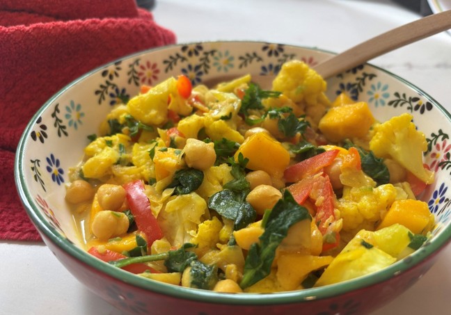 A colorful vegetable curry with chickpeas, cauliflower, spinach, red bell pepper, and chunks of yellow squash in a decorative bowl, with a wooden spoon and a red cloth nearby.