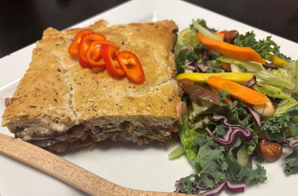 A square piece of savory pie topped with sliced red peppers is served on a white plate next to a mixed green salad with kale, carrots, purple cabbage, and nuts. A wooden fork rests on the plate.