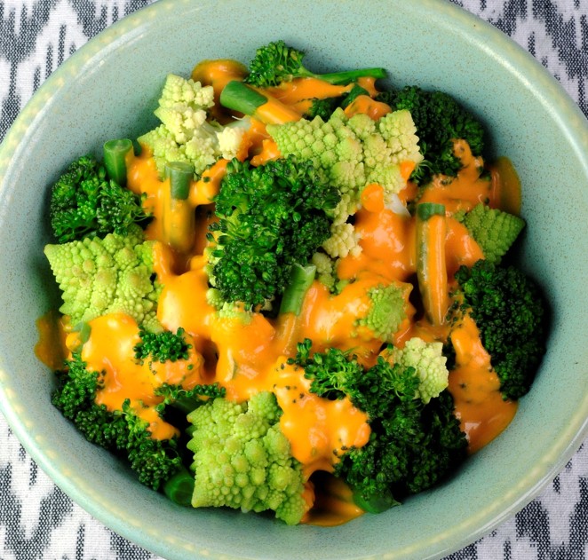A bowl filled with broccoli and Romanesco florets, topped with a generous drizzle of bright orange cheese sauce, sits on a patterned cloth background.