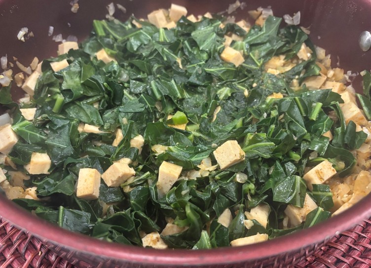 A pan filled with sautéed collard greens, diced tofu, chopped onions, and spices, with the ingredients evenly mixed and cooked.