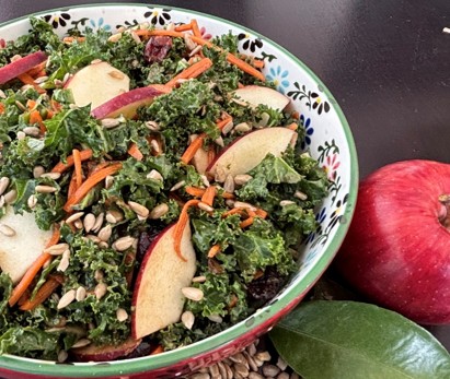 A colorful salad in a patterned bowl featuring kale, sliced apples, shredded carrots, sunflower seeds, and dried cranberries, with a whole red apple and green leaf beside the bowl.