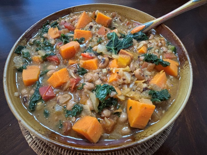A bowl of hearty stew containing chunks of orange sweet potato, leafy greens, tomatoes, beans, and other vegetables, with a spoon resting in the bowl on a wooden table.