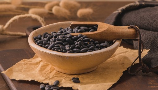 A ceramic bowl filled with black beans sits on a piece of brown parchment paper. A wooden scoop rests on top of the beans, and a burlap sack is visible in the background.