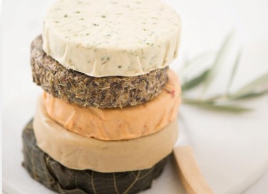 A stack of four round, gourmet cheeses with different colors and textures, arranged on a white surface beside a small wooden knife and a sprig of green leaves.
