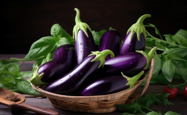 A woven basket filled with shiny, fresh purple eggplants sits on a wooden surface, surrounded by green basil leaves. A few tomatoes and kitchen utensils are visible in the background.