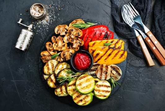 A round platter of grilled vegetables, including zucchini, mushrooms, bell peppers, and onions, arranged around a small bowl of red sauce, with cutlery and a pepper grinder nearby on a dark background.